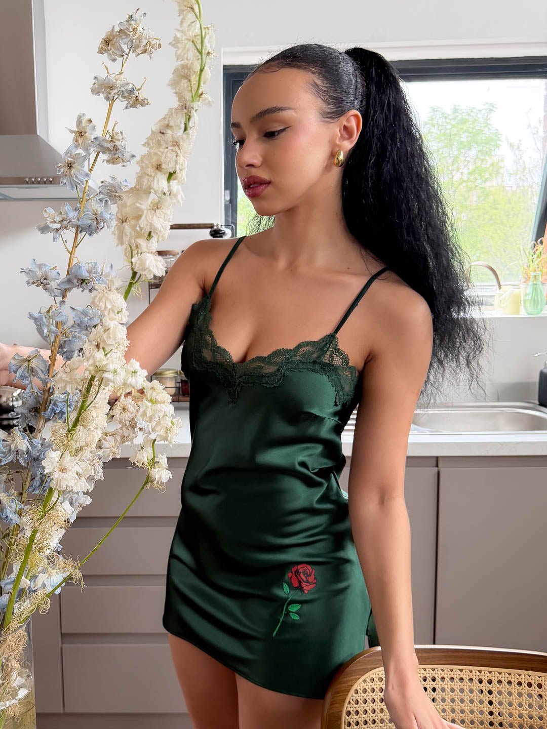 Model wearing an emerald green satin mini chemise featuring a delicate rose print inspired by Italian romance, in a bright modern kitchen.